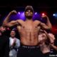 A shirtless Shakur Stevenson flexes both arms on stage during the official weigh-in for his fight against William Zepeda on July 11, 2025, with team members and officials watching behind him.
