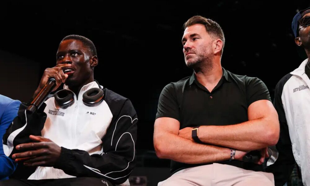 Richardson Hitchins speaks into a microphone during a press event while promoter Eddie Hearn sits beside him with folded arms, listening.